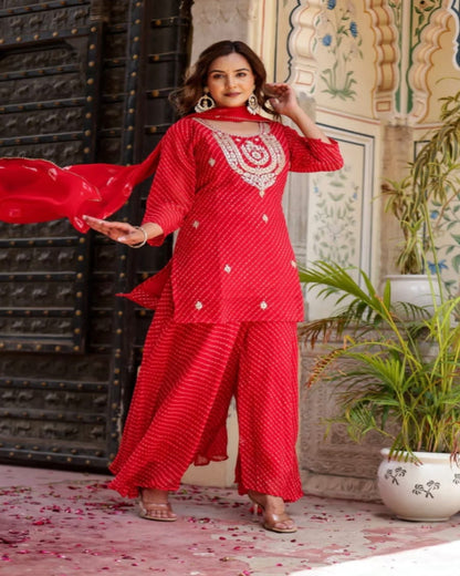 Woman in a red traditional outfit with intricate patterns, standing in an ornate indoor setting.