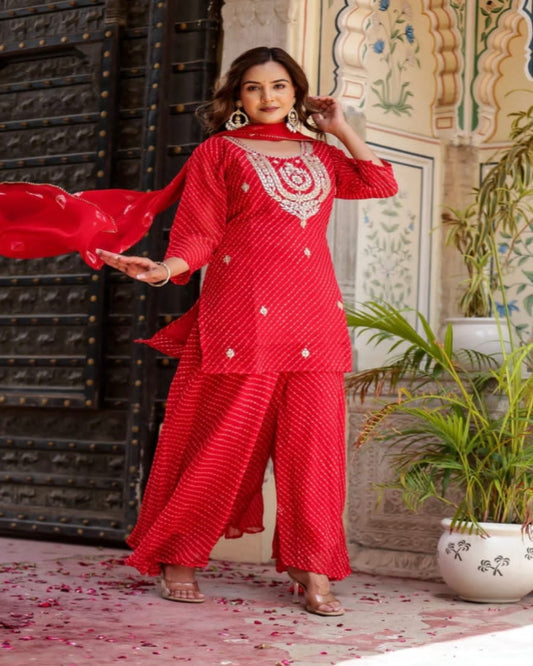 Woman in a red traditional outfit with intricate patterns, standing in an ornate indoor setting.