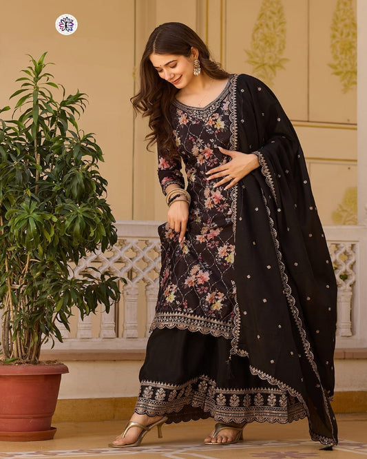 Woman in traditional black and floral outfit standing indoors with a plant and decorative wall in the background