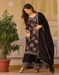 Woman in traditional black and floral outfit standing indoors with a plant and decorative wall in the background
