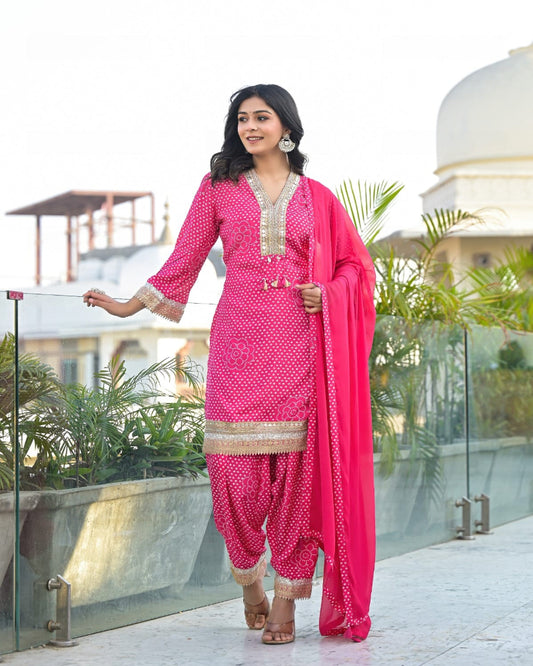 Woman in a bright pink traditional outfit with gold patterns standing on a rooftop with greenery and architectural elements in the background.