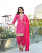 Woman in a bright pink traditional outfit with gold patterns standing on a rooftop with greenery and architectural elements in the background.